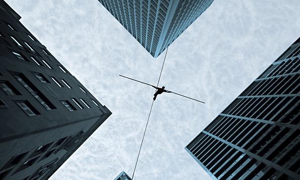 view looking up at a person walking a tightrope between tall buildings 